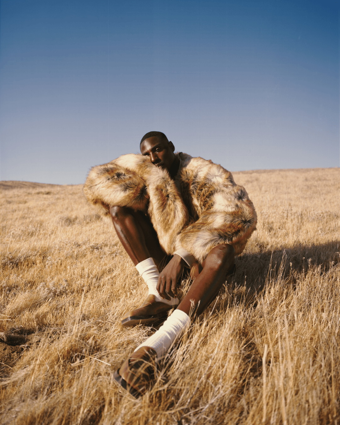 A man in a fur coat sitting in a field of golden grass under a blue sky.