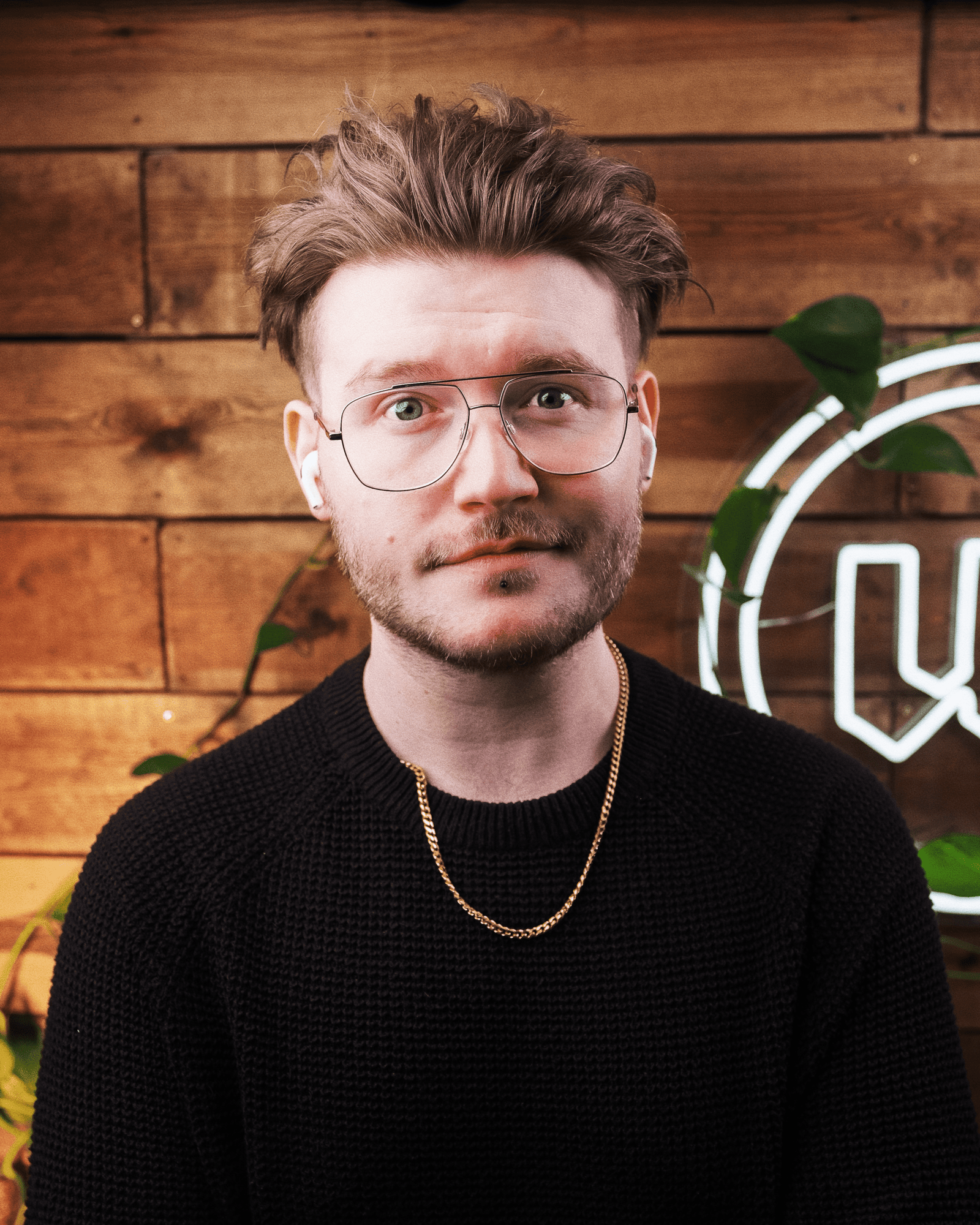 Retrato del diseñador de la marca, Will Paterson, con gafas y un jersey negro, de pie frente a una pared de madera con plantas y un discreto letrero de neón al fondo.