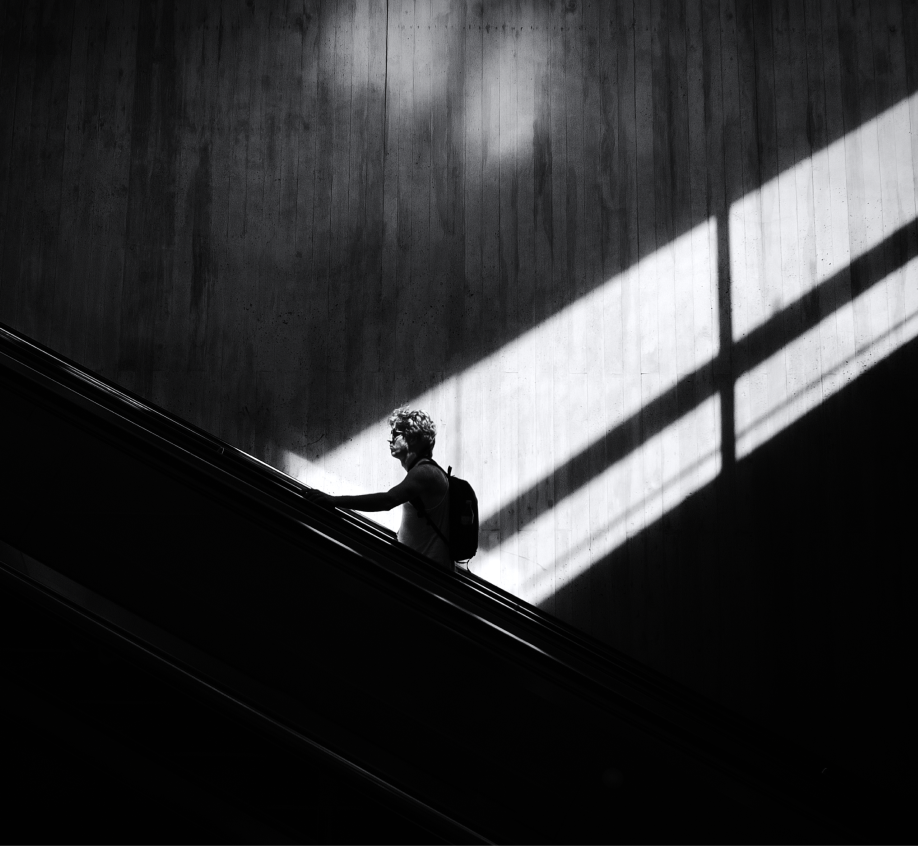 Black and white photo of a person with a backpack riding an escalator, with dramatic sunlight and shadow patterns on the wall behind them.