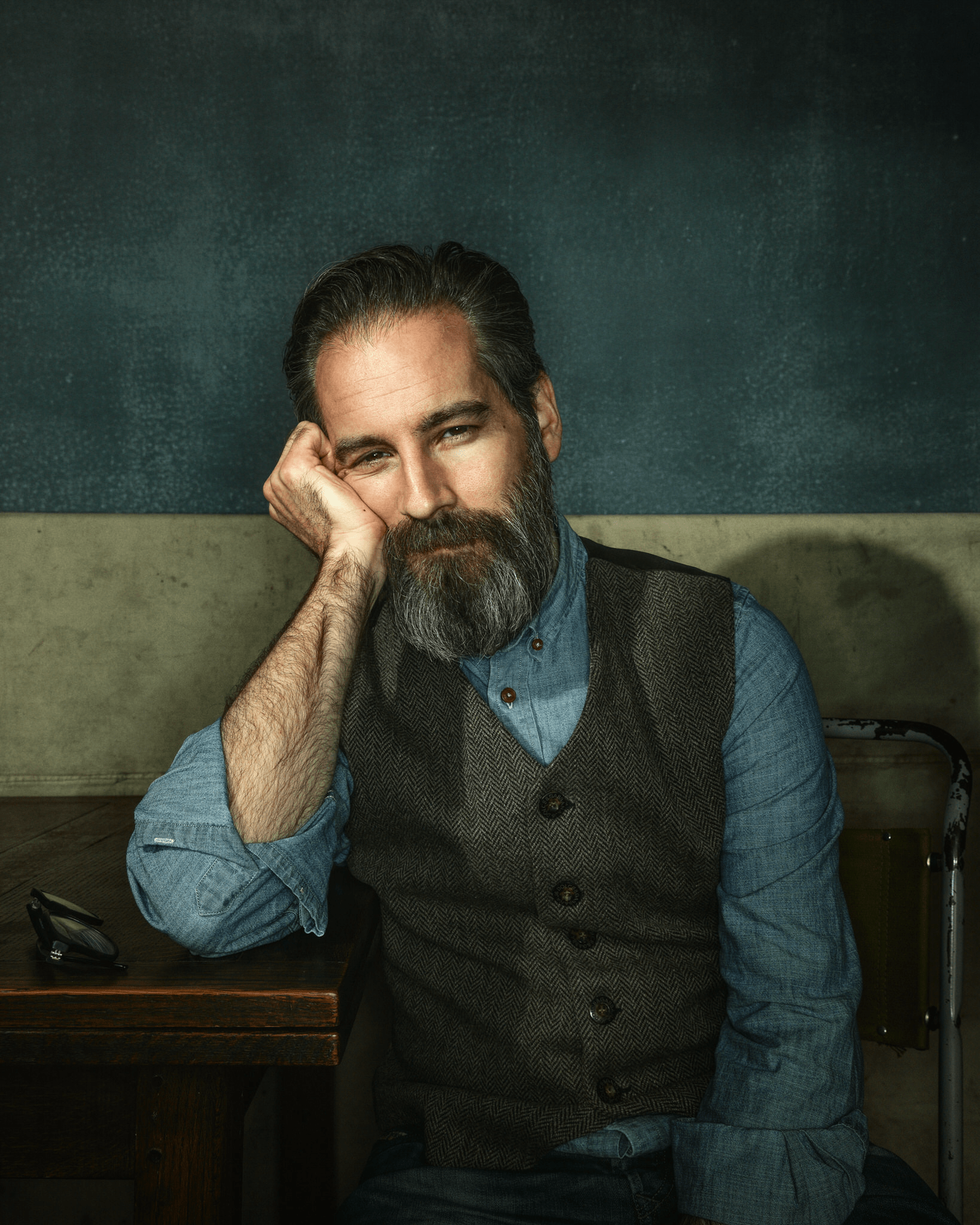 Self-portrait of photographer Ivan Weiss seated beside a wooden table, wearing a blue shirt and gray waistcoat, with his head resting on one hand.