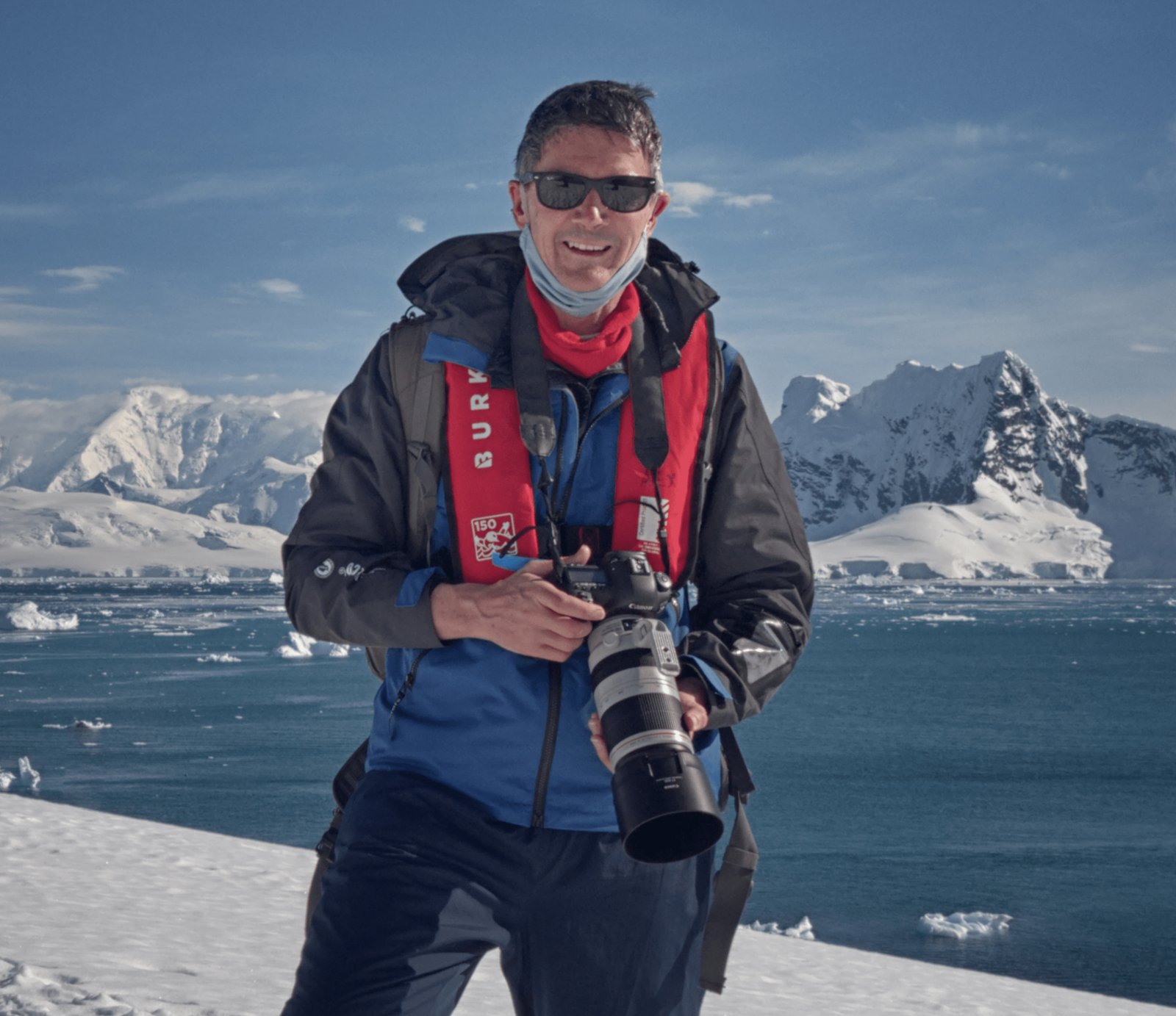 Portrait of photographer Paul Harrison standing on a snowy Antarctic shoreline, holding a camera with snow-covered mountains and floating icebergs behind him.