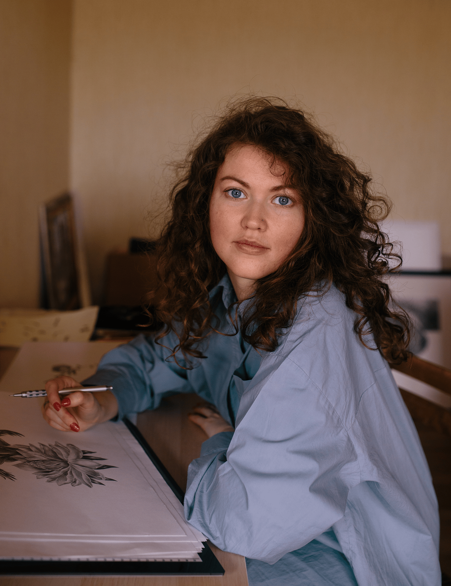 Portrait of illustrator Mary Maka holding a pencil and looking at the camera while seated at a desk.