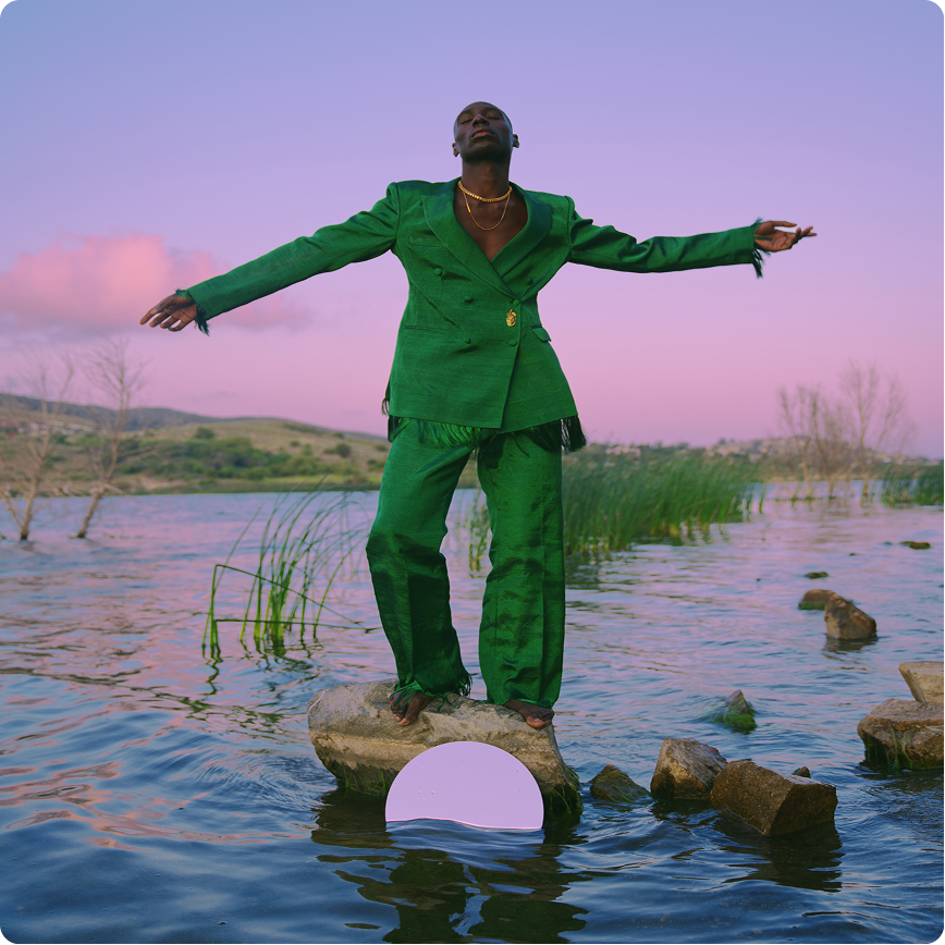 Person in a bright green suit standing barefoot on a rock in shallow water, arms outstretched, with a pink and purple sunset sky.