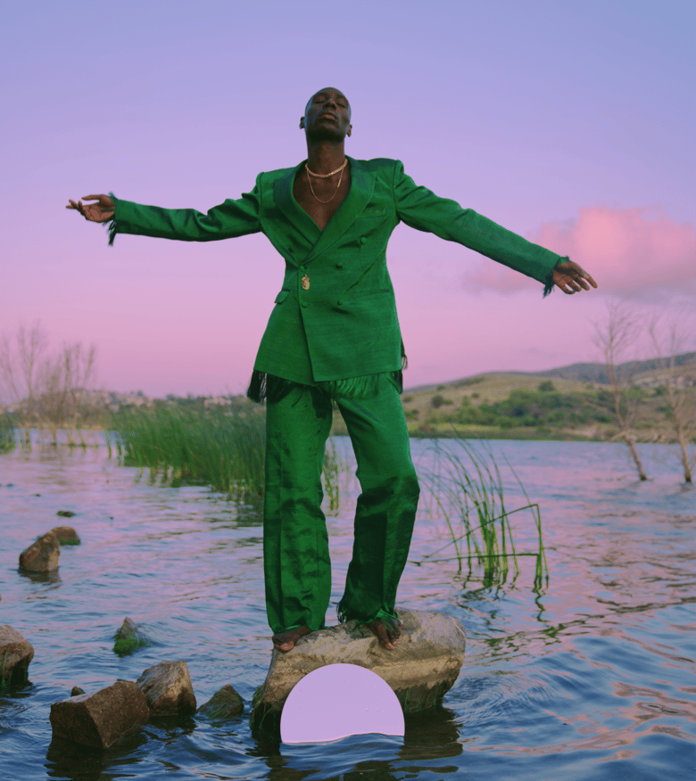Person in a green suit standing on a rock in a calm lake at sunset with arms outstretched